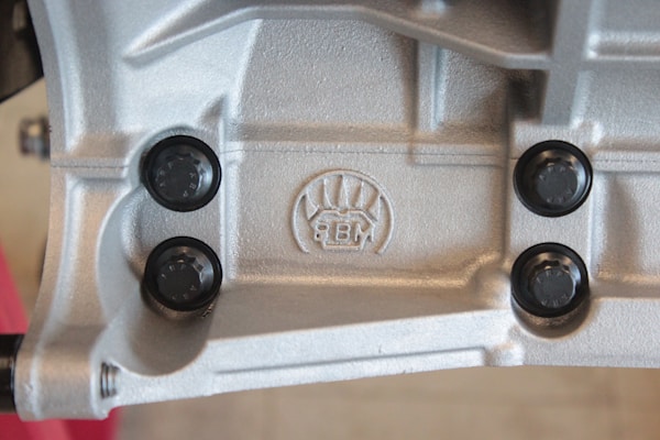 A close-up view of a metallic surface with detailed textures and patterns. Four black, round fasteners are symmetrically placed around an embossed logo featuring stylized graphics and the letters 'BM'. The metallic surface has a silver-gray color and appears industrial in nature.