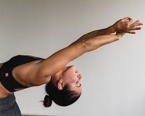 A woman is performing a yoga stretch, leaning backward with arms extended overhead. She is wearing a black sports bra and has her hair tied back. The pose highlights the muscles in her arms and upper body, demonstrating strength and flexibility.