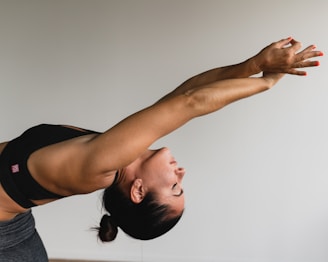 A woman is performing a yoga stretch, leaning backward with arms extended overhead. She is wearing a black sports bra and has her hair tied back. The pose highlights the muscles in her arms and upper body, demonstrating strength and flexibility.