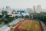 An aerial view of a sports complex with a running track surrounding a grassy field, set against the background of a cityscape. The city's skyline features a mix of tall residential buildings and shorter commercial structures. There are trees and green areas surrounding the sports facility.