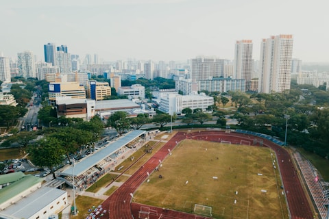 An aerial view of a sports complex with a running track surrounding a grassy field, set against the background of a cityscape. The city's skyline features a mix of tall residential buildings and shorter commercial structures. There are trees and green areas surrounding the sports facility.