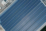 An aerial view of an array of solar panels arranged in parallel rows on a rooftop. The panels have a uniform grid pattern and reflect sunlight. The surrounding area includes metal structures and rooftop machinery, indicating an industrial setting.