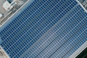 An aerial view of an array of solar panels arranged in parallel rows on a rooftop. The panels have a uniform grid pattern and reflect sunlight. The surrounding area includes metal structures and rooftop machinery, indicating an industrial setting.
