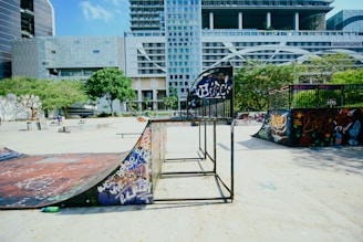 A vibrant urban skate park featuring ramps, graffiti, and modern architecture in the background. Trees and greenery add a touch of nature to the concrete setting, with people casually enjoying the space.