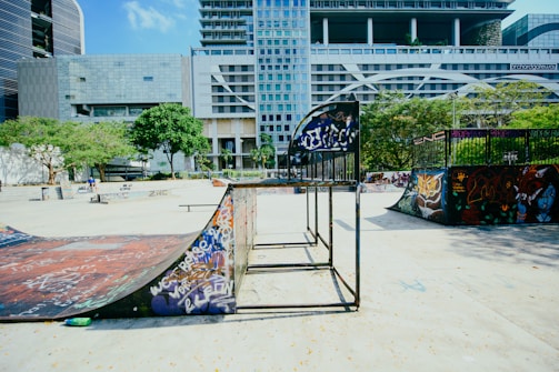 A vibrant urban skate park featuring ramps, graffiti, and modern architecture in the background. Trees and greenery add a touch of nature to the concrete setting, with people casually enjoying the space.