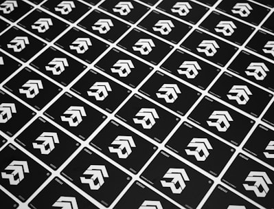 A grid of identical black cards with a white geometric logo, arranged diagonally. The cards have text at the bottom corners, and the logo consists of three cascading arrow-like shapes.