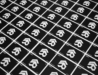 A grid of identical black cards with a white geometric logo, arranged diagonally. The cards have text at the bottom corners, and the logo consists of three cascading arrow-like shapes.