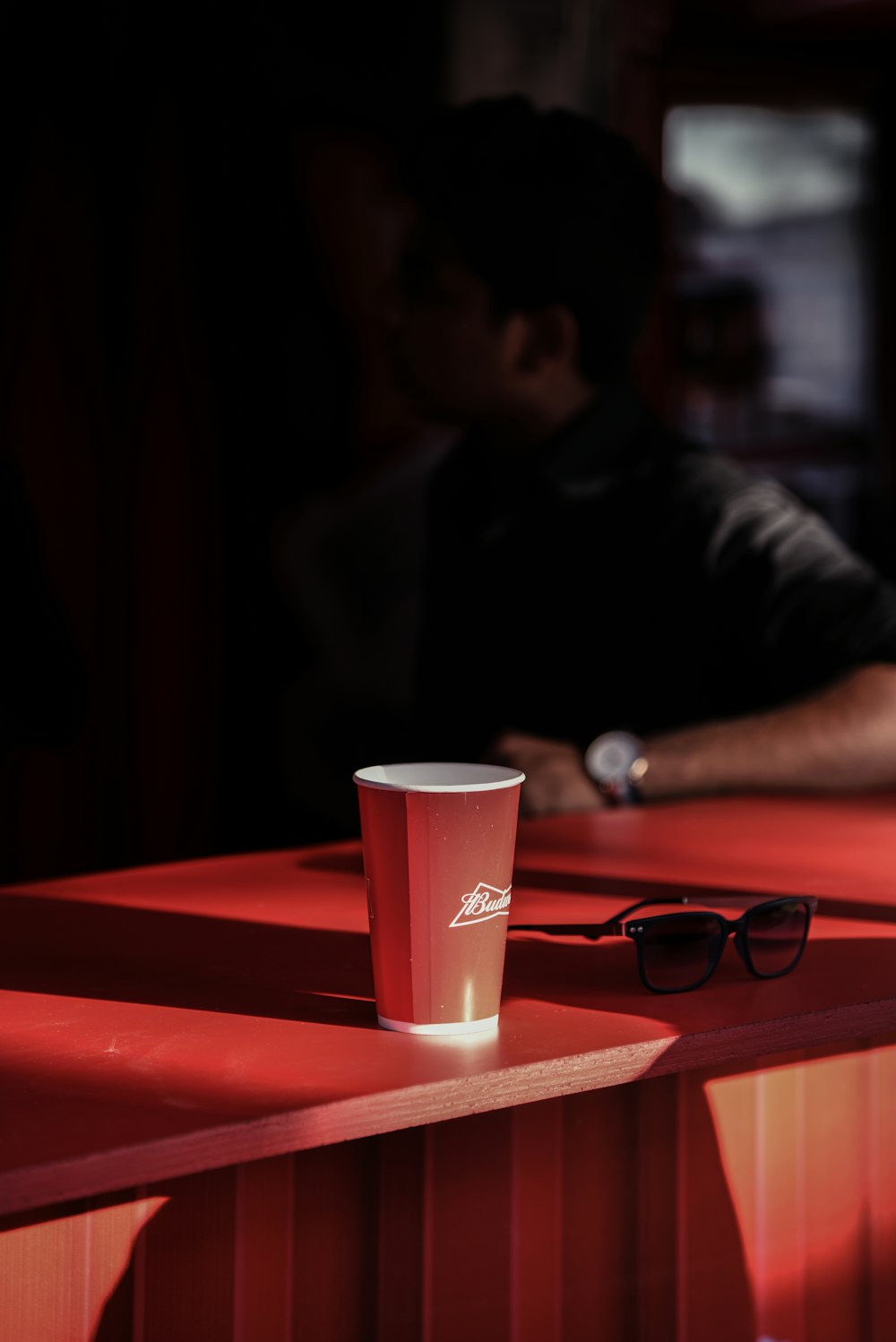 man sitting beside red and white cup next to black framed sunglasses