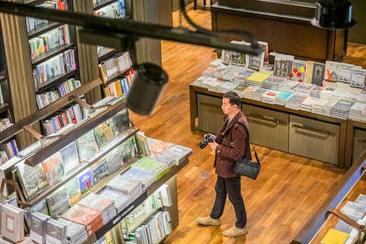 A person holds a camera while standing in a bookstore, surrounded by shelves filled with a variety of books. There are large tables displaying books with colorful covers. The wooden floor adds warmth to the environment.