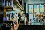 A cozy coffee shop interior with shelves neatly lined with various bags of coffee beans and merchandise. In the foreground, there are clear glass coffee brewers on a dark wooden counter. The bright natural light filtering through large windows illuminates the space, creating a warm and inviting atmosphere.