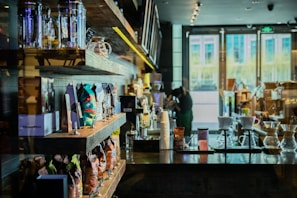 A cozy coffee shop interior with shelves neatly lined with various bags of coffee beans and merchandise. In the foreground, there are clear glass coffee brewers on a dark wooden counter. The bright natural light filtering through large windows illuminates the space, creating a warm and inviting atmosphere.
