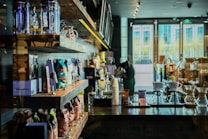 A cozy coffee shop interior with shelves neatly lined with various bags of coffee beans and merchandise. In the foreground, there are clear glass coffee brewers on a dark wooden counter. The bright natural light filtering through large windows illuminates the space, creating a warm and inviting atmosphere.