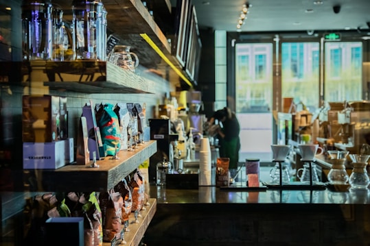 A cozy coffee shop interior with shelves neatly lined with various bags of coffee beans and merchandise. In the foreground, there are clear glass coffee brewers on a dark wooden counter. The bright natural light filtering through large windows illuminates the space, creating a warm and inviting atmosphere.
