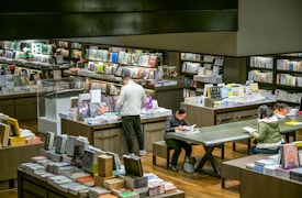 A bookstore interior with a variety of books on shelves and display tables. People are browsing or reading books, with one person standing at a table and others seated. The room is well-lit, creating a cozy and inviting atmosphere.