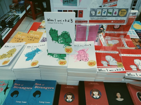 A display of various books on a table, featuring multiple copies of a series titled 'When I was a kid' with different volume numbers and cover illustrations in green, pink, blue, and yellow. Each book has a 20% discount sticker. Other visible titles include 'Sea of Strangers' by Lang Leav, and there are book cover images of various styles visible in the background.