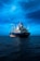 A large blue cargo ship is prominently positioned in the middle of a calm sea under a vast, somewhat cloudy sky. The ship has several antennas and cranes on deck, with the name 'XINRUI' visible on its hull.