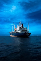 A large blue cargo ship is prominently positioned in the middle of a calm sea under a vast, somewhat cloudy sky. The ship has several antennas and cranes on deck, with the name 'XINRUI' visible on its hull.