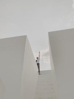 A person stands at the top of a set of white stairs between two large white walls, reaching one hand upwards towards the sky. The sky is partly cloudy, creating a serene and minimalistic scene.