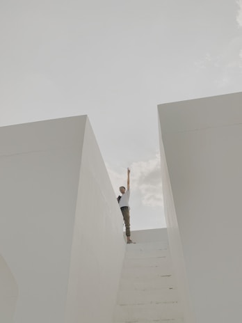 A person stands at the top of a set of white stairs between two large white walls, reaching one hand upwards towards the sky. The sky is partly cloudy, creating a serene and minimalistic scene.
