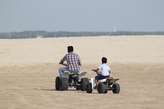 Two people are riding all-terrain vehicles (ATVs) on a sandy landscape. The person on the left is larger and likely an adult, wearing a checkered shirt and jeans, riding a bigger ATV. The person on the right is smaller, possibly a child, wearing a light shirt and shorts, riding a smaller ATV. The background shows a vast sandy expanse under a clear sky.