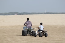 Two people are riding all-terrain vehicles (ATVs) on a sandy landscape. The person on the left is larger and likely an adult, wearing a checkered shirt and jeans, riding a bigger ATV. The person on the right is smaller, possibly a child, wearing a light shirt and shorts, riding a smaller ATV. The background shows a vast sandy expanse under a clear sky.