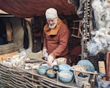 A person with a gray beard and a woolen cap appears to be engaged in a traditional craft or trade within a rustic setting. They are dressed in medieval-style clothing and are surrounded by natural materials and handmade objects, such as wooden tools, clay bowls, and wool. The setting includes woven wooden fencing and a tent-like background, suggesting a historical reenactment or market scene.