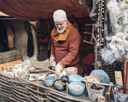 A person with a gray beard and a woolen cap appears to be engaged in a traditional craft or trade within a rustic setting. They are dressed in medieval-style clothing and are surrounded by natural materials and handmade objects, such as wooden tools, clay bowls, and wool. The setting includes woven wooden fencing and a tent-like background, suggesting a historical reenactment or market scene.