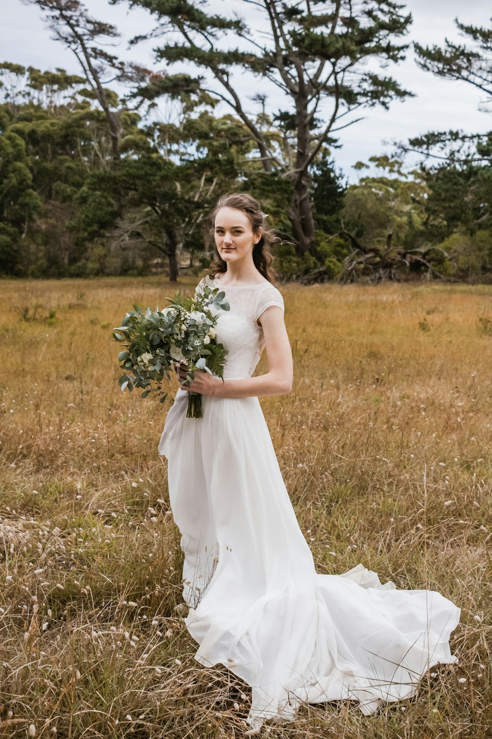 woman wearing white wedding dress standing on pasture while holding bouquet during daytime
