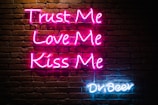 Neon sign displays the phrases 'Trust Me, Love Me, Kiss Me' in pink lettering and 'Dr Beer' in blue lettering on a dark brick wall background.