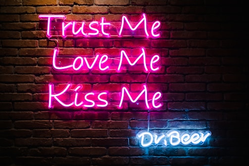 Neon sign displays the phrases 'Trust Me, Love Me, Kiss Me' in pink lettering and 'Dr Beer' in blue lettering on a dark brick wall background.