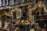 An assortment of ornate Venetian masks is displayed behind a shop window. The elaborate designs feature gold and black hues, with intricate patterns and feathers. The reflection of an old building adds a layer of complexity to the scene.