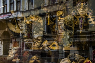 An assortment of ornate Venetian masks is displayed behind a shop window. The elaborate designs feature gold and black hues, with intricate patterns and feathers. The reflection of an old building adds a layer of complexity to the scene.