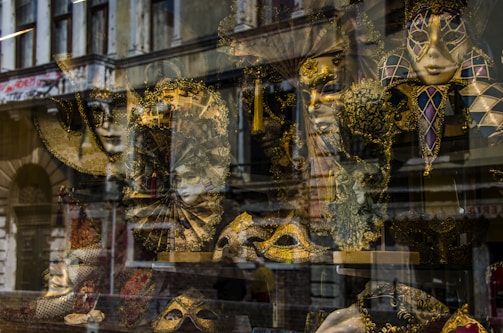 An assortment of ornate Venetian masks is displayed behind a shop window. The elaborate designs feature gold and black hues, with intricate patterns and feathers. The reflection of an old building adds a layer of complexity to the scene.