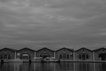 A row of five identical industrial-style warehouses is situated by the water. The buildings have arched windows and brick exteriors. A few boats are docked along the waterfront, and there are some umbrellas and seating areas near the buildings. The sky is overcast with clouds, creating a somber atmosphere.