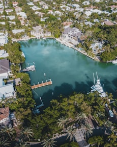An aerial view of an upscale residential area featuring a large, serene body of water surrounded by lush greenery and palm trees. Elegant houses with swimming pools line the water's edge, some with private docks extending into the water. Several boats are moored nearby, and the area is densely populated with trees and vegetation.