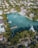 An aerial view of an upscale residential area featuring a large, serene body of water surrounded by lush greenery and palm trees. Elegant houses with swimming pools line the water's edge, some with private docks extending into the water. Several boats are moored nearby, and the area is densely populated with trees and vegetation.
