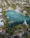 An aerial view of an upscale residential area featuring a large, serene body of water surrounded by lush greenery and palm trees. Elegant houses with swimming pools line the water's edge, some with private docks extending into the water. Several boats are moored nearby, and the area is densely populated with trees and vegetation.