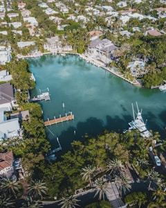 An aerial view of an upscale residential area featuring a large, serene body of water surrounded by lush greenery and palm trees. Elegant houses with swimming pools line the water's edge, some with private docks extending into the water. Several boats are moored nearby, and the area is densely populated with trees and vegetation.