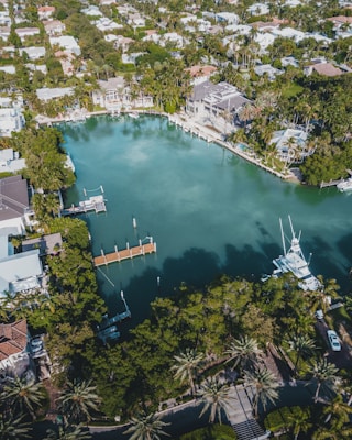 An aerial view of an upscale residential area featuring a large, serene body of water surrounded by lush greenery and palm trees. Elegant houses with swimming pools line the water's edge, some with private docks extending into the water. Several boats are moored nearby, and the area is densely populated with trees and vegetation.