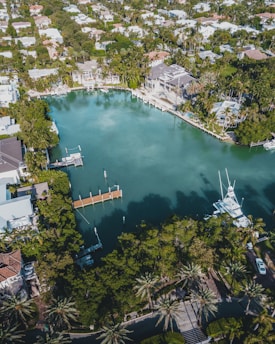 An aerial view of an upscale residential area featuring a large, serene body of water surrounded by lush greenery and palm trees. Elegant houses with swimming pools line the water's edge, some with private docks extending into the water. Several boats are moored nearby, and the area is densely populated with trees and vegetation.