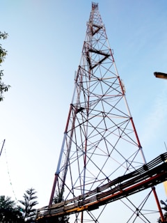 A tall communication tower constructed with a lattice of metal beams is set against a clear blue sky. The structure features a series of crisscrossing patterns and is anchored with various cables. Trees and other foliage are visible at the bottom of the image.