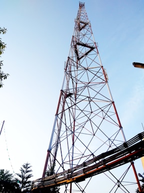 A tall communication tower constructed with a lattice of metal beams is set against a clear blue sky. The structure features a series of crisscrossing patterns and is anchored with various cables. Trees and other foliage are visible at the bottom of the image.