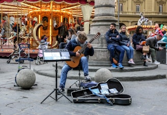 A street musician performs on a guitar in a lively urban setting. A group of people sit on steps, listening and observing. Behind them, a vintage carousel with decorative horses is brightly lit, creating a warm, vibrant backdrop. Nearby, various street objects like stones and strollers are visible, adding to the dynamic city atmosphere.