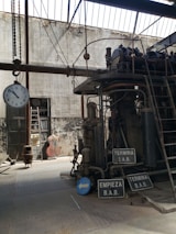An industrial setting featuring a large, complex mechanical machine with various pipes and gears. The environment appears aged, with a high ceiling and large windows allowing natural light to cast shadows. A clock is hanging from a chain, and several signs with Spanish text are visible. The background shows a worn wall, a door, and some storage items.