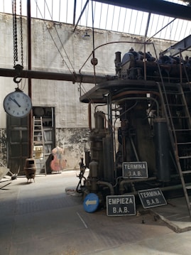 An industrial setting featuring a large, complex mechanical machine with various pipes and gears. The environment appears aged, with a high ceiling and large windows allowing natural light to cast shadows. A clock is hanging from a chain, and several signs with Spanish text are visible. The background shows a worn wall, a door, and some storage items.