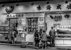 A black and white photograph of an Asian street market storefront. Several people, including two children and two adults, are seen standing and browsing items displayed through a glass window and hanging from hooks. The store has multiple items for sale including packages, jars, and hanging goods. Chinese characters are prominent on the sign above the store and the hanging signs.