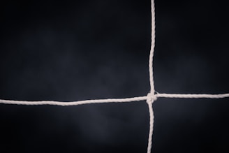 A white rope forms a knot at the intersection of two perpendicular cords against a dark, blurred background. The focus is on the center where the cords intersect.