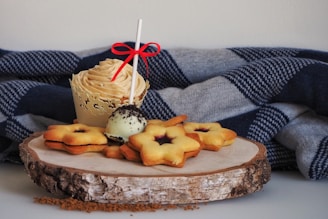 A decorative cupcake with a creamy swirl on top sits beside a group of star-shaped sandwich cookies and a cake pop on a wooden platter. The backdrop features a cozy, folded blanket with a blue and gray checkered pattern, enhancing the cozy and inviting atmosphere.