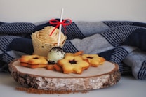 A decorative cupcake with a creamy swirl on top sits beside a group of star-shaped sandwich cookies and a cake pop on a wooden platter. The backdrop features a cozy, folded blanket with a blue and gray checkered pattern, enhancing the cozy and inviting atmosphere.