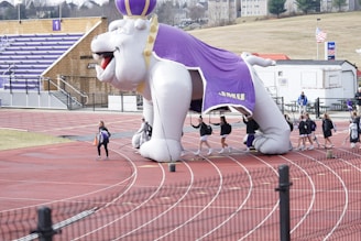 A large inflatable bulldog mascot with a purple cape and crown is positioned on a running track. Several individuals are walking underneath the inflatable, indicating a sports team. The background features bleachers with purple seating and a sign with the number one.